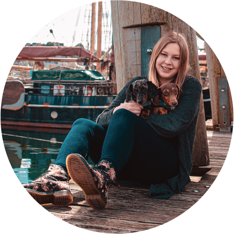 A woman with long hair sits on a wooden dock, holding two dachshund puppies. She is smiling and wearing patterned boots. Behind her are tall masts and the hull of a boat, suggesting a marina setting.